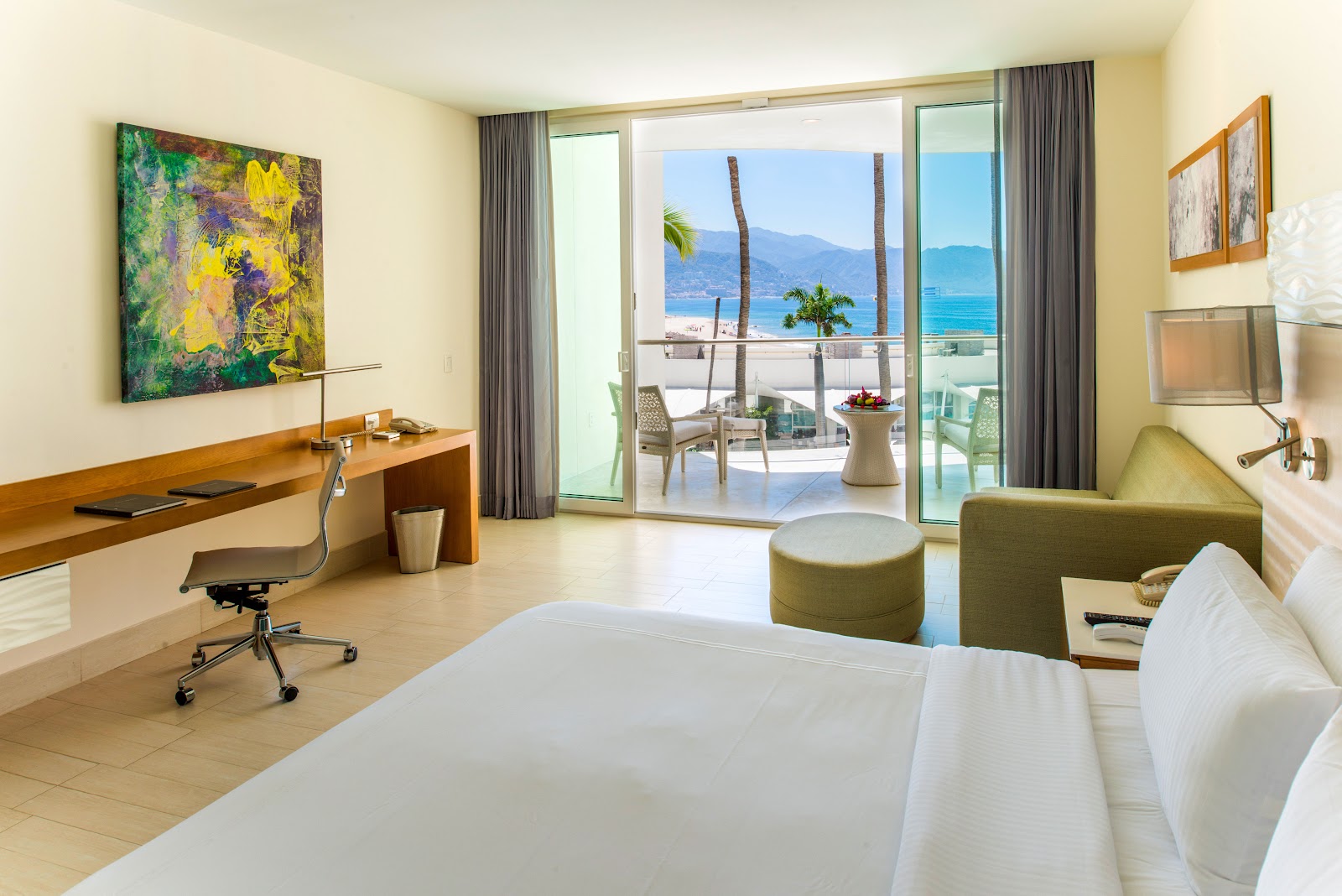 Resort room with bed, work desk, and sliding glass doors opening to a balcony overlooking palm trees and blue ocean water with mountains beyond.
