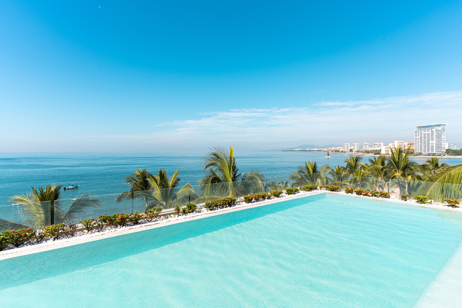Turquoise infinity pool overlooking the Pacific Ocean with palm trees, white sand, and coastal resort buildings in the distance.