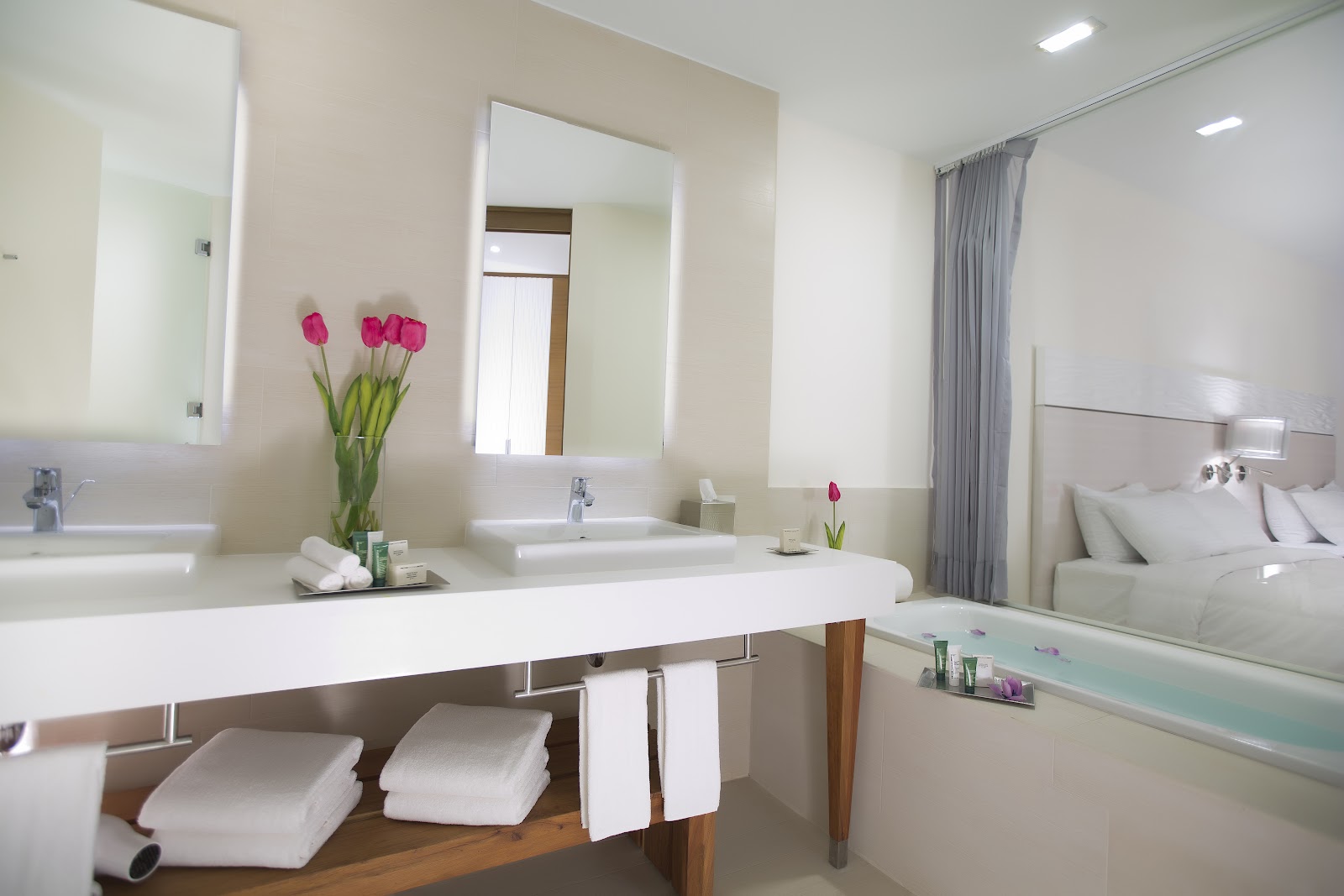 Modern bathroom with dual sinks, lighted mirrors, pink tulips, white towels, and soaking tub visible at Krystal Grand Puerto Vallarta.