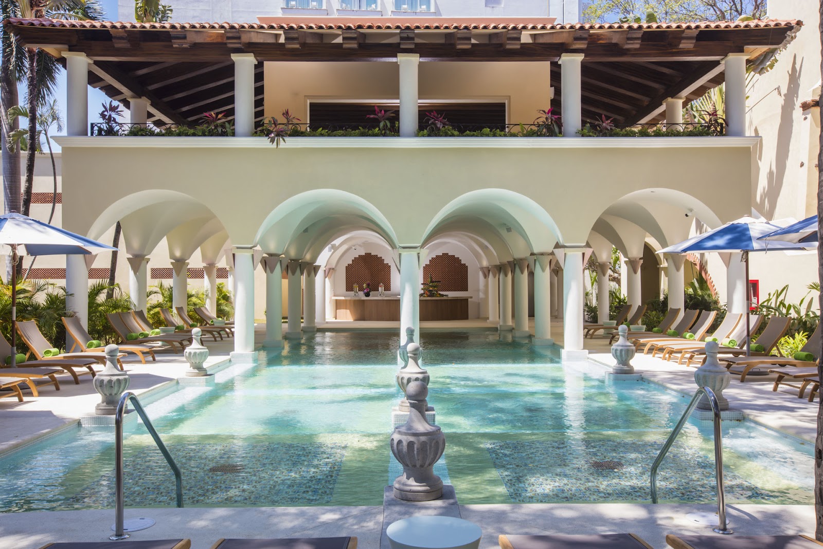 Indoor pool with turquoise water under arched colonnades at Krystal Grand Puerto Vallarta, flanked by lounge chairs and blue umbrellas.