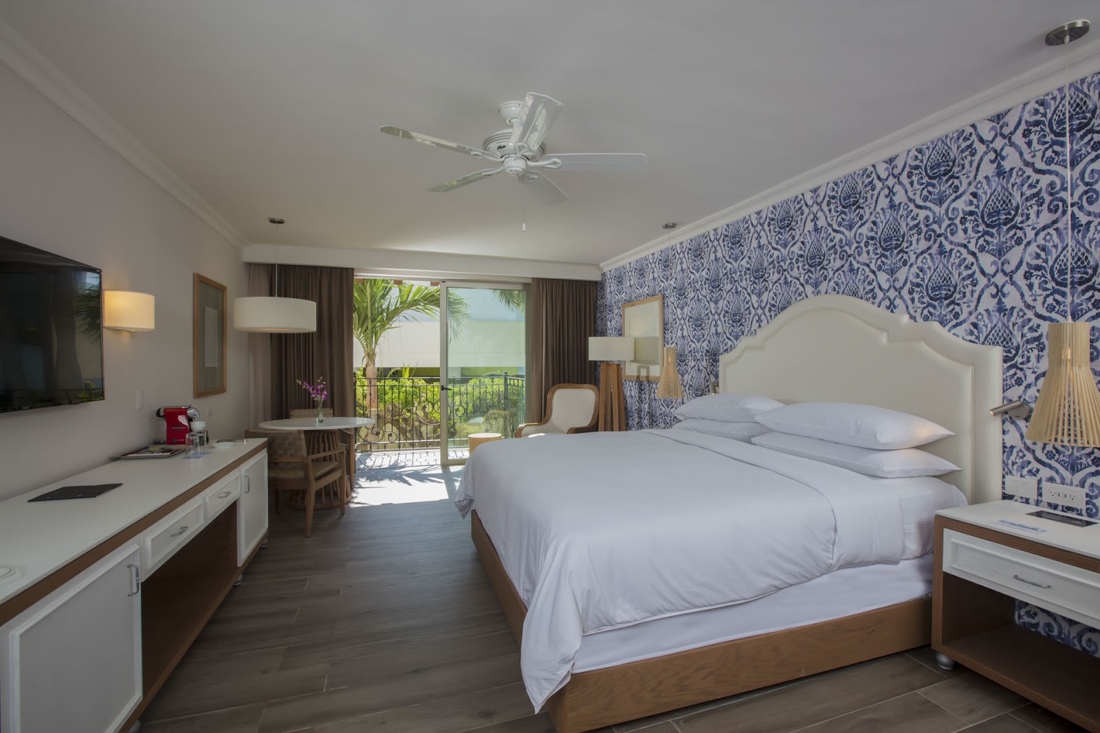 Resort guestroom with queen bed, blue and white patterned accent wall, desk, and ocean-view balcony with palm trees visible.