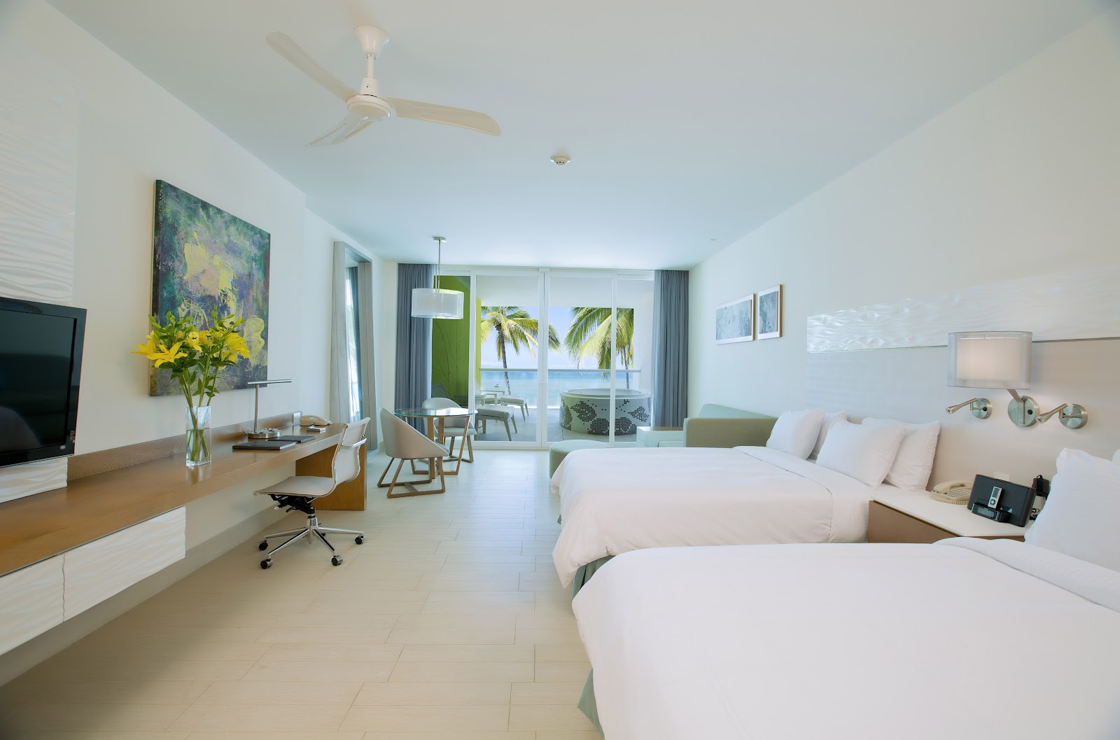 Modern resort room with two beds, work desk, flat-screen TV, and ocean view through balcony doors with palm trees visible.