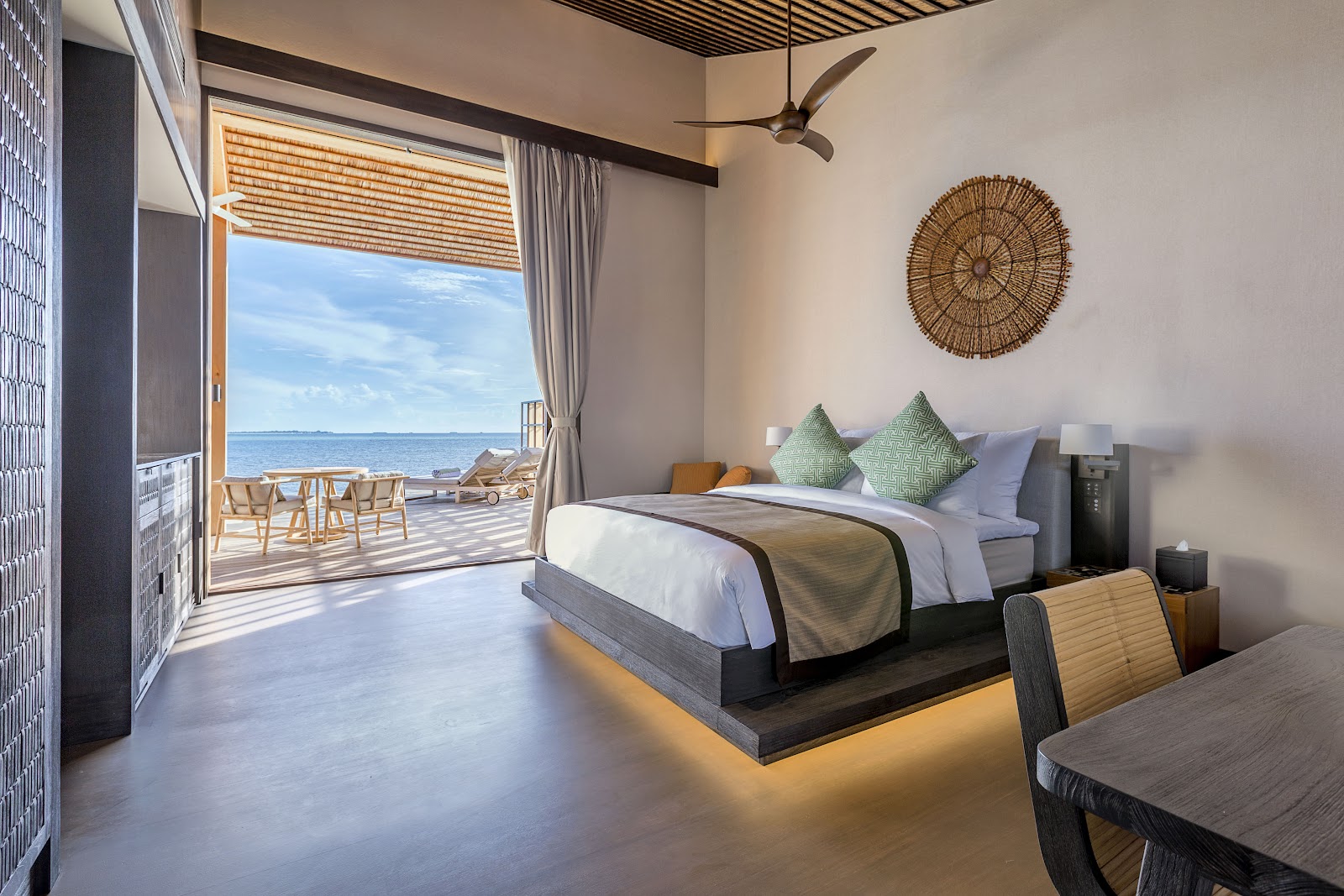 Beachfront bedroom at Kudadoo Maldives Private Island with ocean view from open terrace, bed with green pillows, woven wall art, and wooden deck furniture.