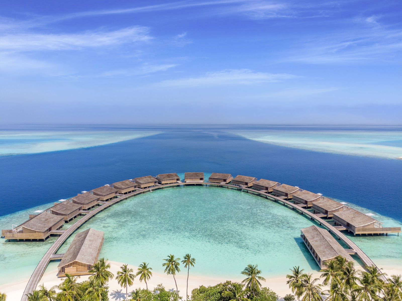 Aerial view of Kudadoo Maldives Private Island showing overwater bungalows arranged in a semicircle around a turquoise lagoon with white sand beach and palm trees.