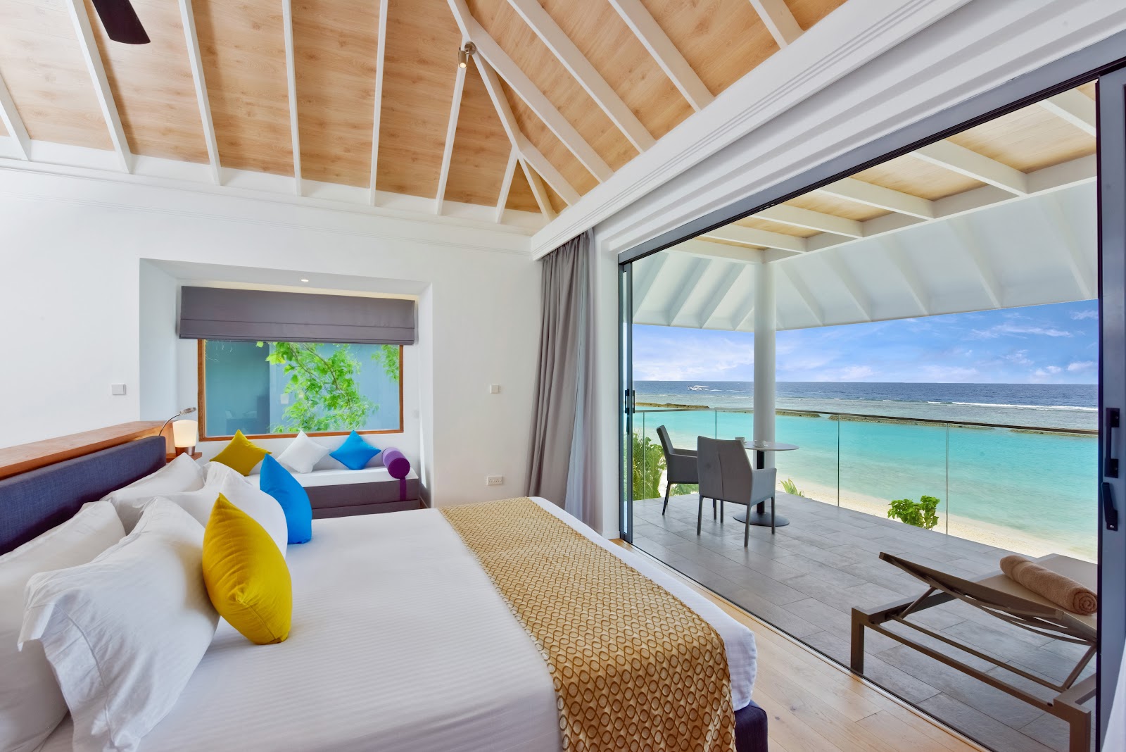 Beachfront bedroom at Kuramathi Island Resort with king bed, colorful pillows, wooden vaulted ceiling, and glass doors opening to terrace overlooking turquoise lagoon.