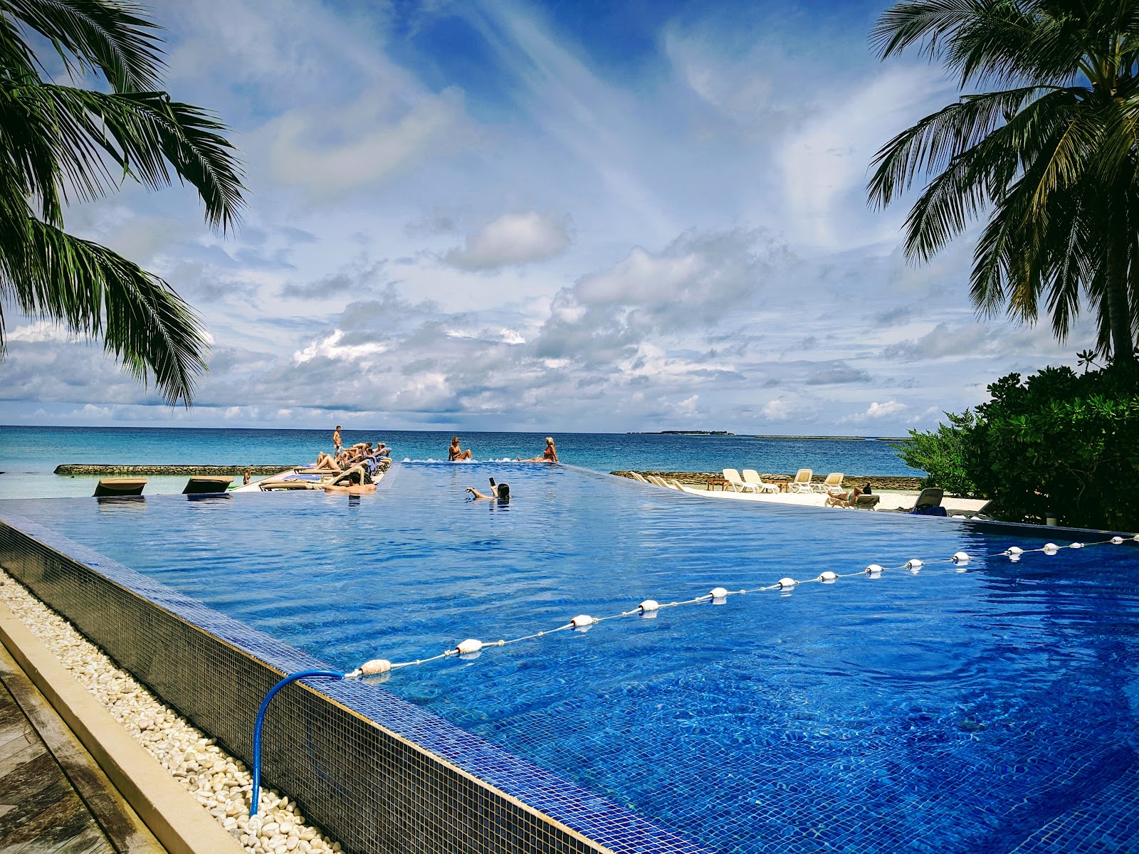 Tiled lagoon pool with rope divider at Kuramathi Island Resort, guests swimming in shallow water beyond, palm trees and ocean horizon under cloudy sky.