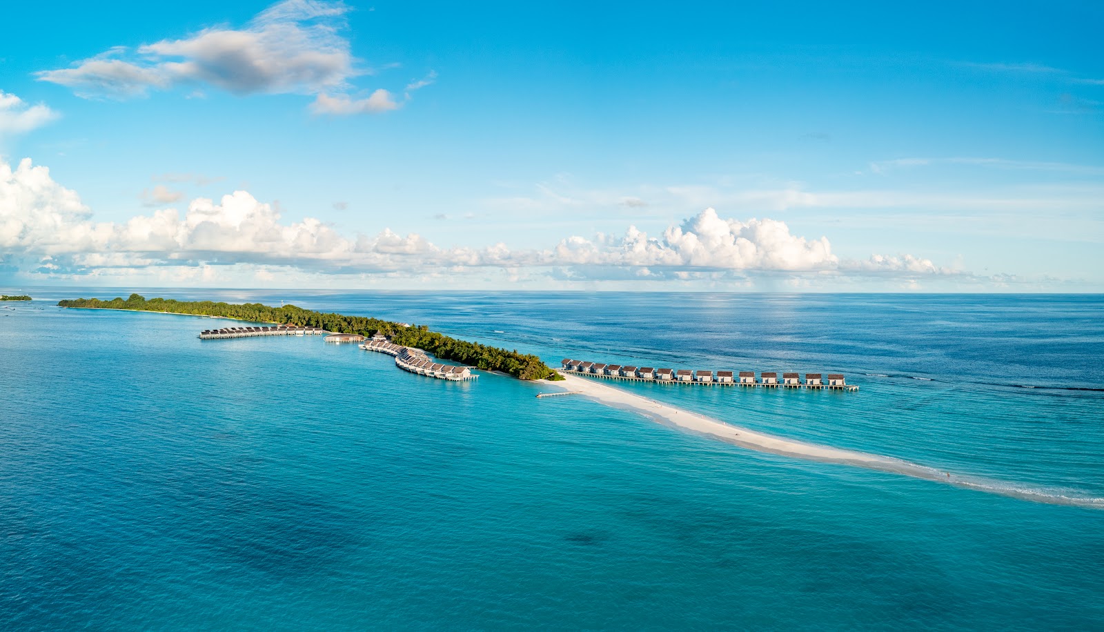 Aerial view of Kuramathi Island Resort with overwater bungalows lining a white sand beach and tropical vegetation under a bright blue sky.