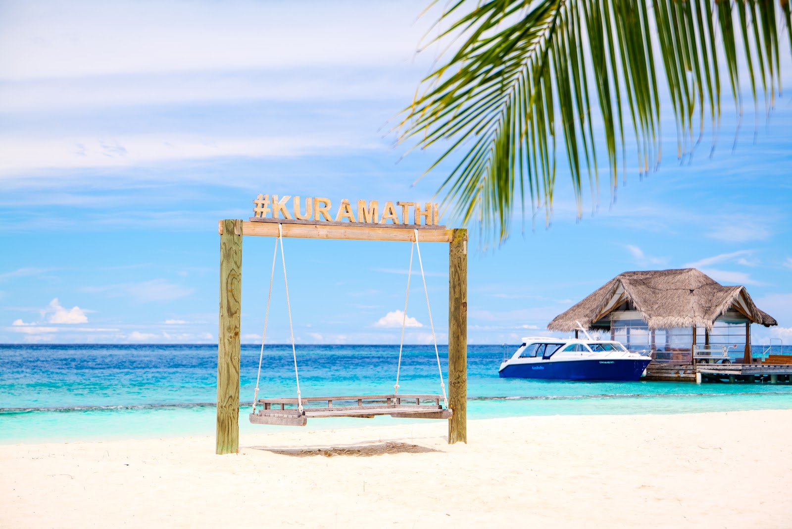 Wooden swing with Kuramathi sign on white sand beach, with turquoise water, boat, and thatched bungalows beyond.