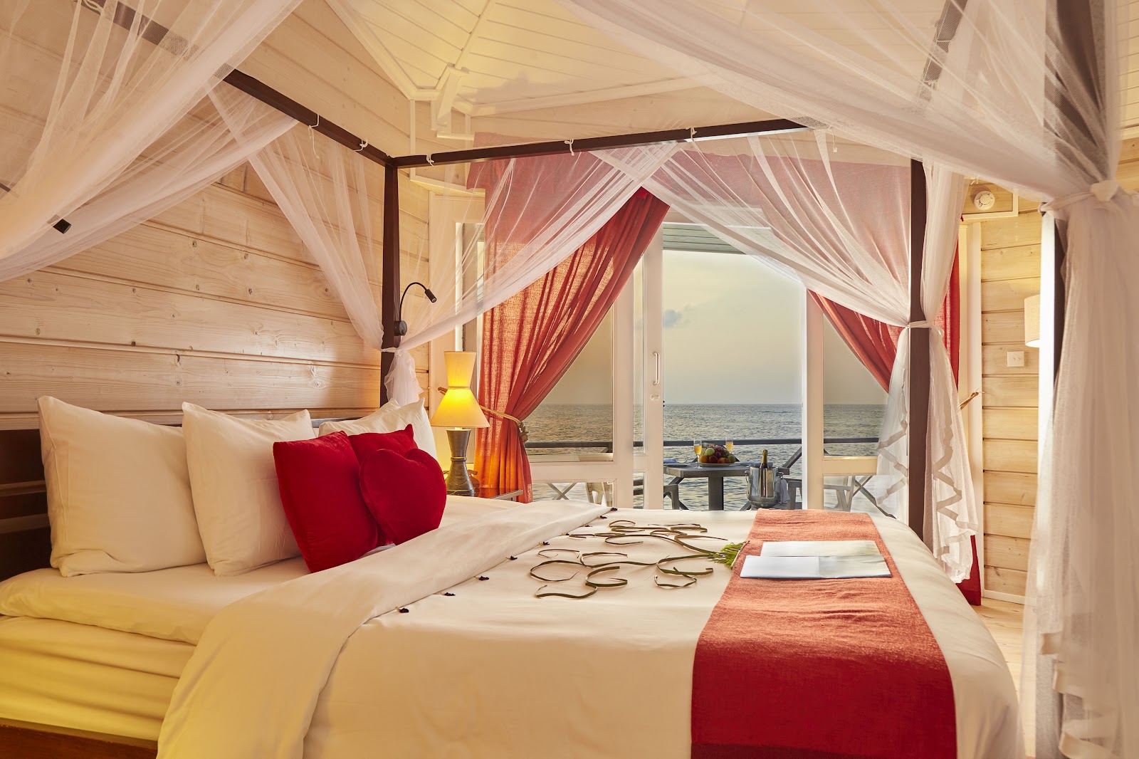 Beachfront suite with four-poster canopy bed draped in sheer curtains, red accent pillows, and ocean view through open glass doors at Kuredu Island Resort & Spa.