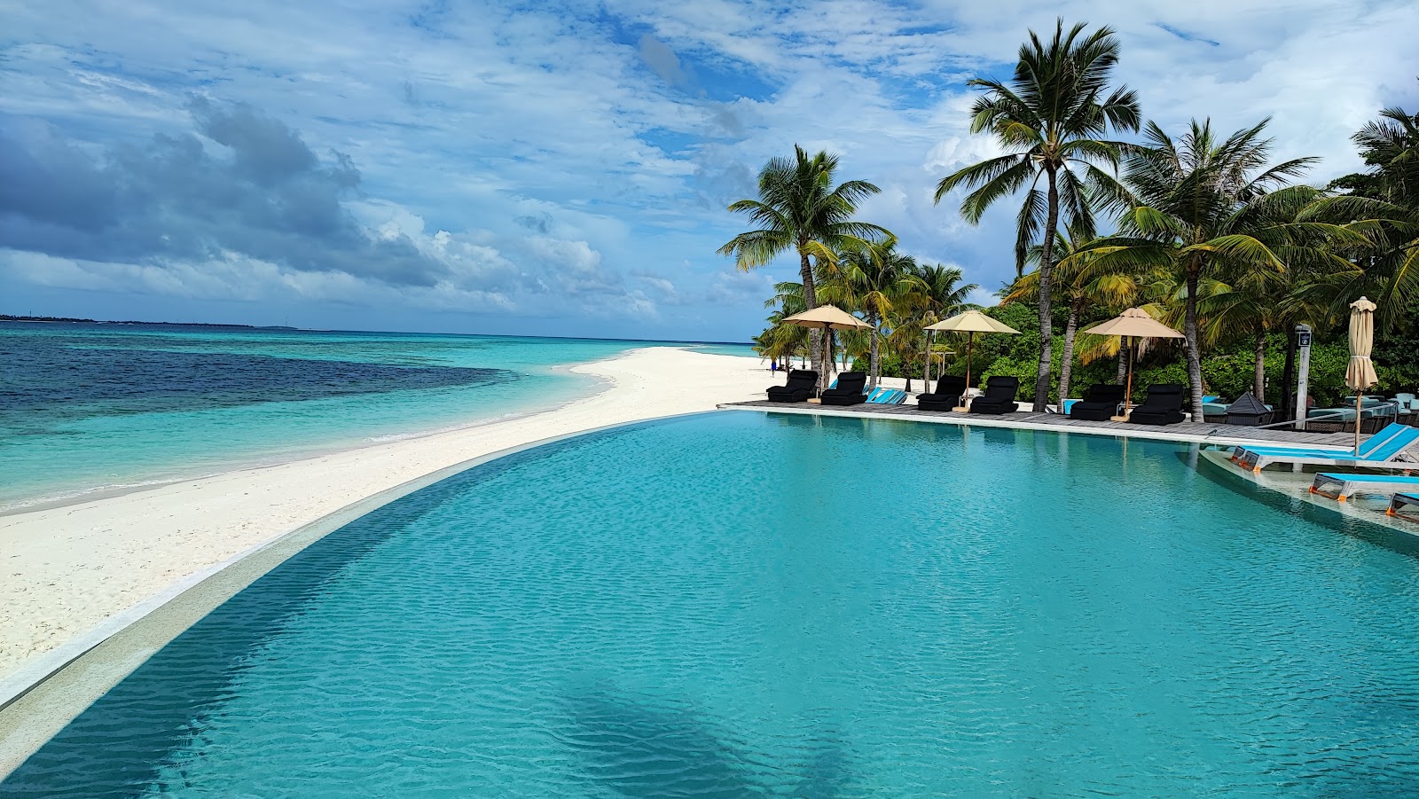 Infinity pool overlooking white sand beach and turquoise ocean at Kuredu Island Resort & Spa, with palm trees and lounge umbrellas.