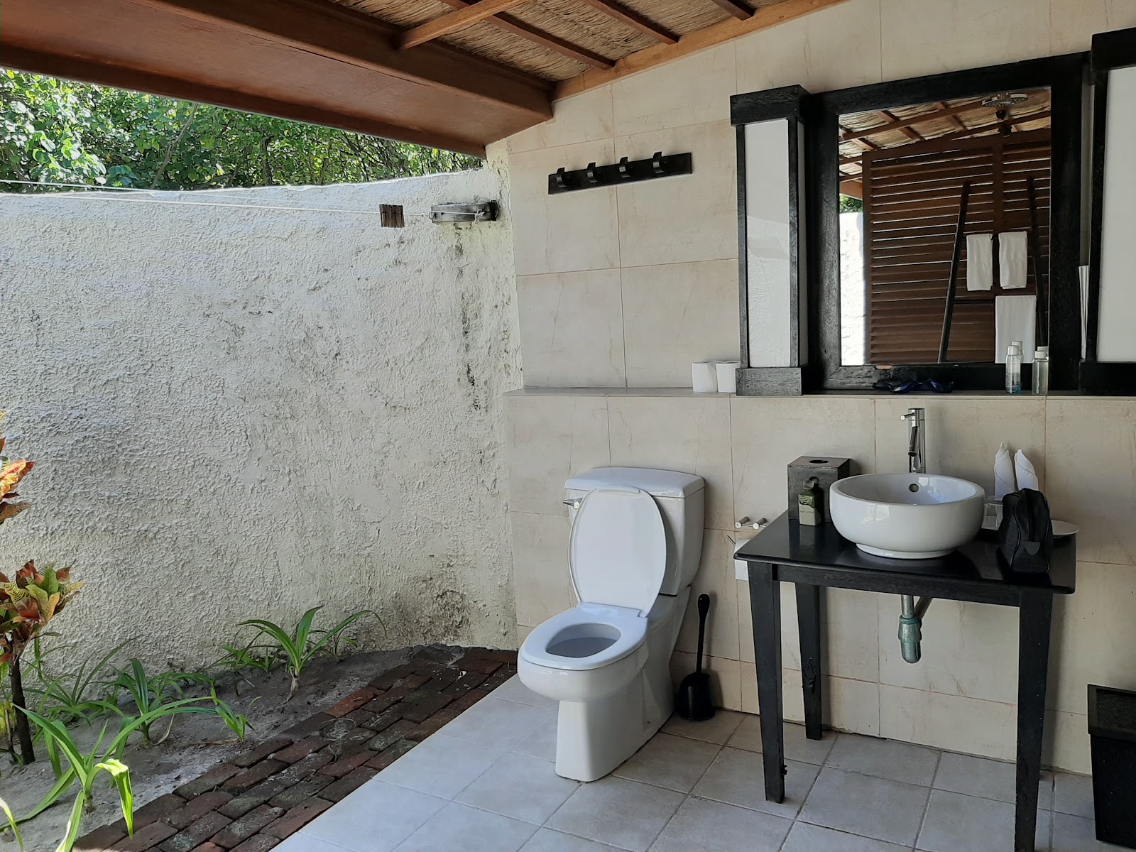 Open-air bathroom with toilet, round white sink on black vanity, large mirror, and outdoor shower wall surrounded by vegetation.
