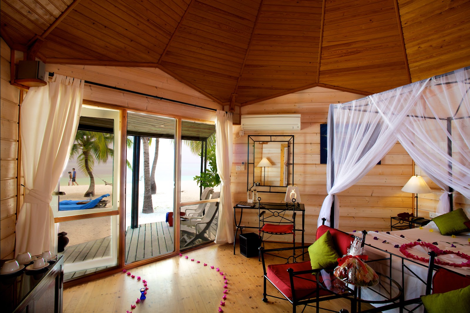 Wooden overwater bungalow interior with canopy bed, open doors to beach, palm trees, and ocean view at Kuredu Island Resort & Spa.