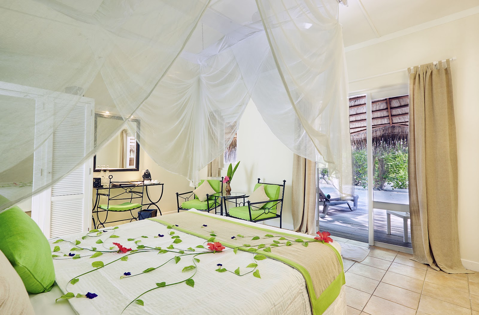 Resort bedroom with sheer canopy draping, green bedding decorated with flower petals, and glass doors opening to a tropical deck.