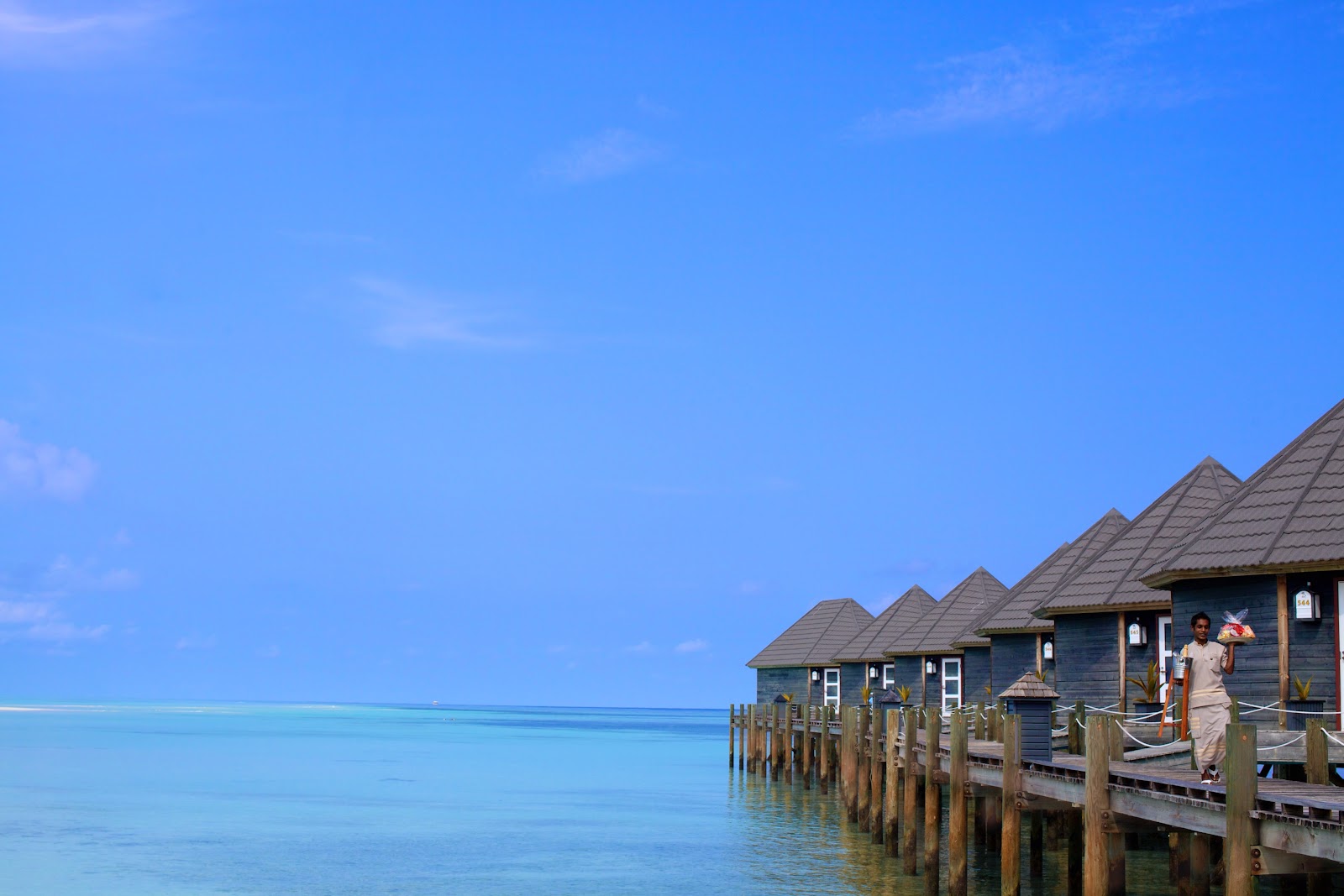 Overwater bungalows with peaked roofs line a wooden pier at Kuredu Island Resort & Spa, with turquoise lagoon and clear blue sky beyond.