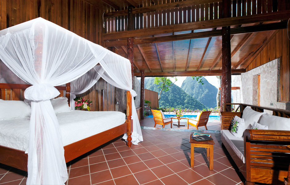 Spacious wooden bungalow with canopy bed, lounge seating, and open-air veranda overlooking Pitons and pool at Ladera Resort.