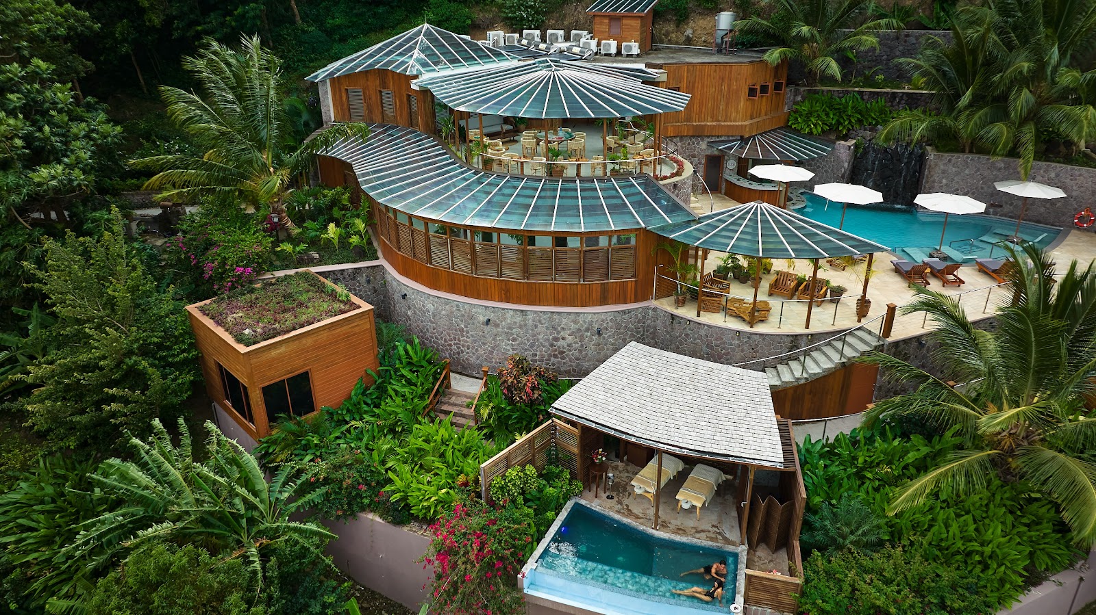 Aerial view of Ladera Resort's wooden bungalows with metal cone roofs nestled among lush tropical vegetation, featuring pools and lounging areas.