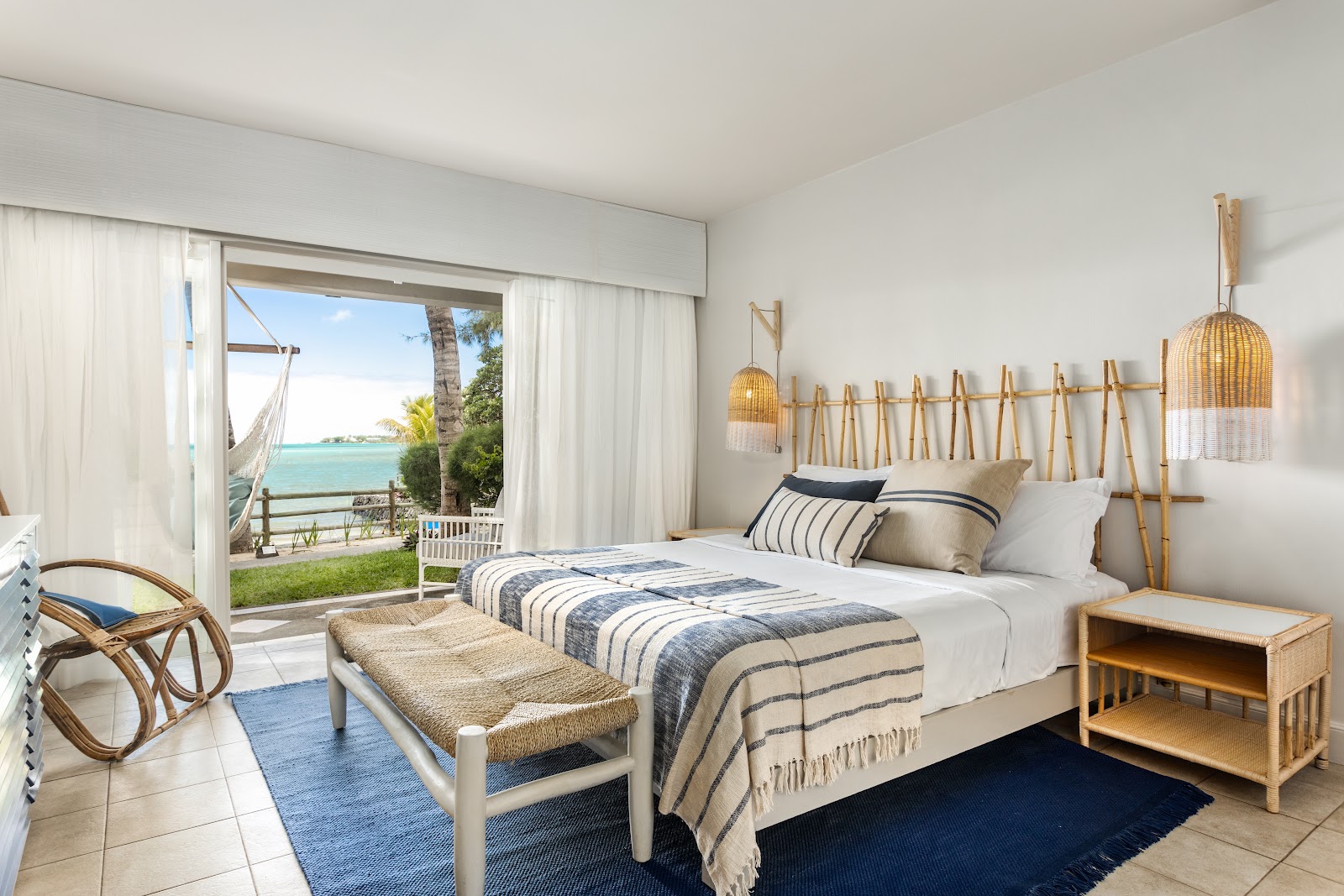Bright bedroom with king bed, woven pendant lights, and ocean view through open glass doors to terrace with hammock and turquoise water.