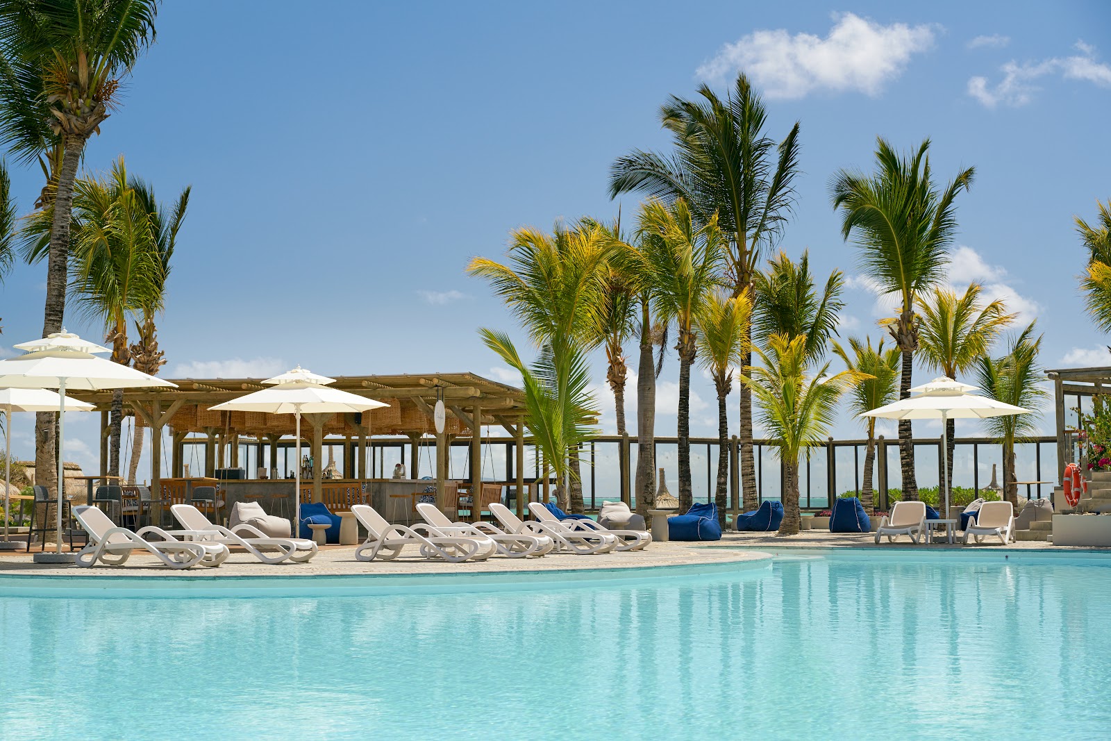 Turquoise pool with white loungers and wooden cabanas shaded by cream umbrellas, surrounded by tall palm trees under blue sky.
