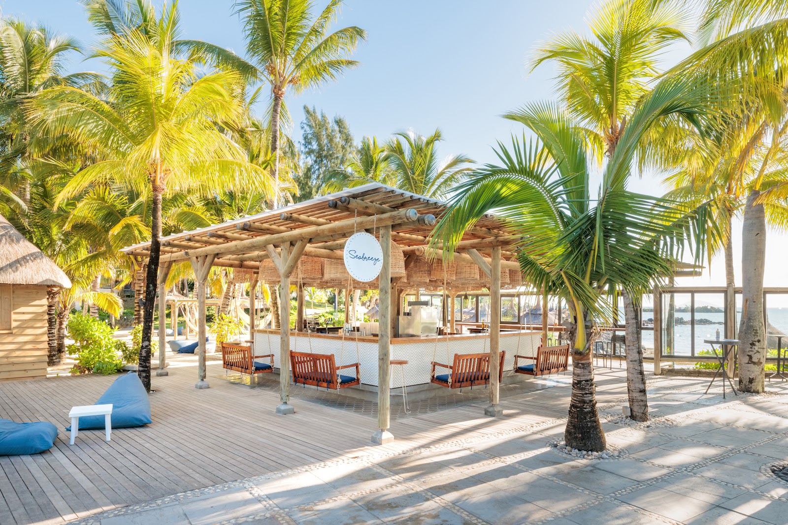 Wooden pergola shelter with brick restaurant building surrounded by palm trees and lounge chairs on a waterfront deck at Lagoon Attitude Hotel.