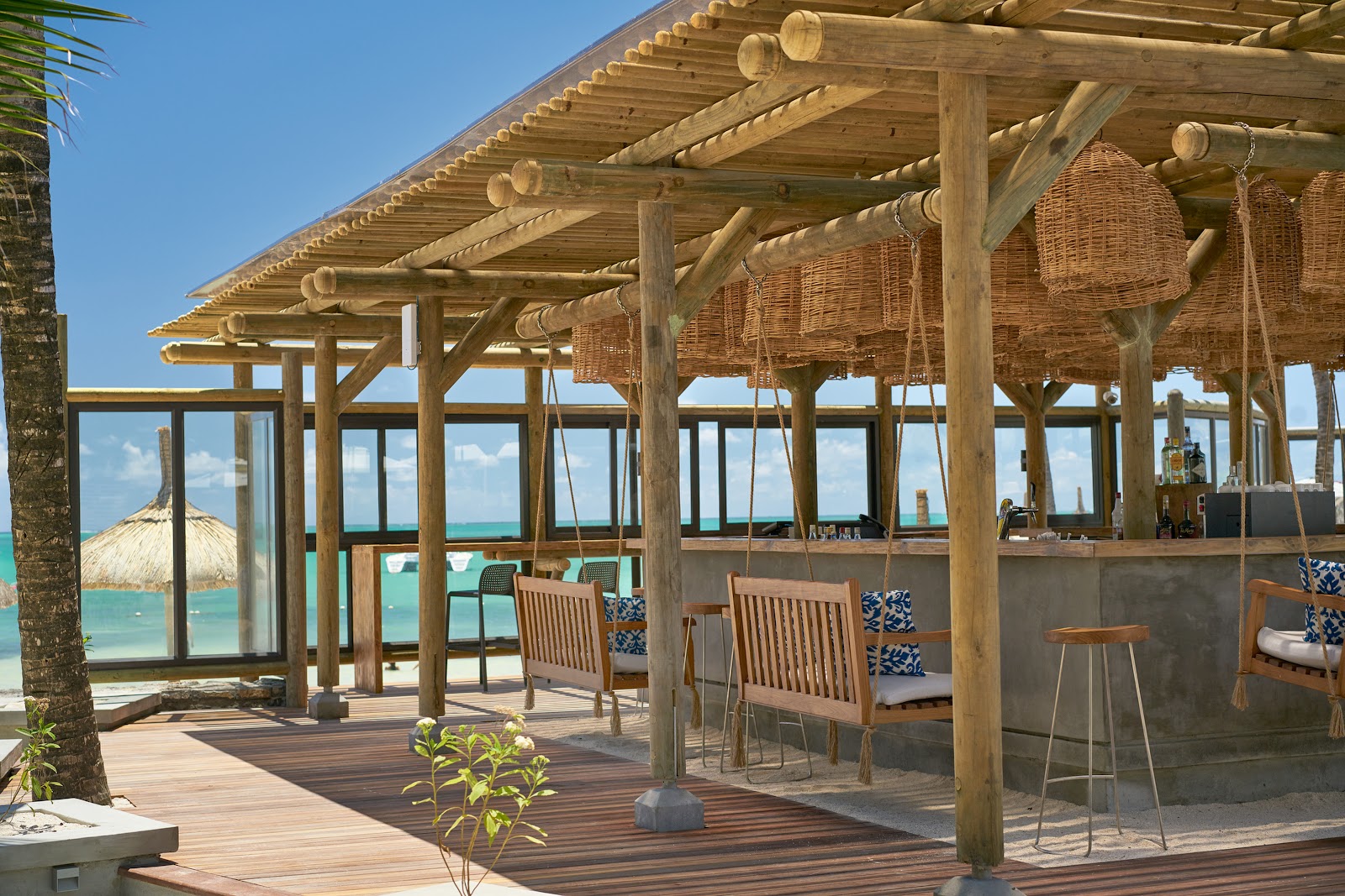 Wooden beam pergola with dining chairs and bar seating overlooking turquoise lagoon water and thatched-roof structures at Lagoon Attitude Hotel.