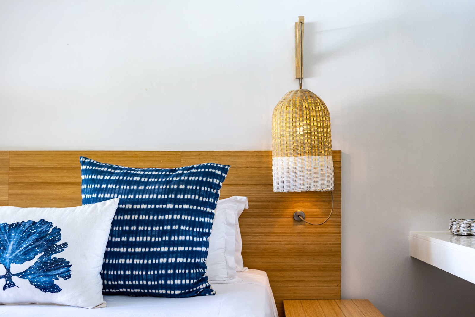 Bed with blue patterned pillows and white sheets against a wooden headboard, with a woven pendant lamp and white wall behind.