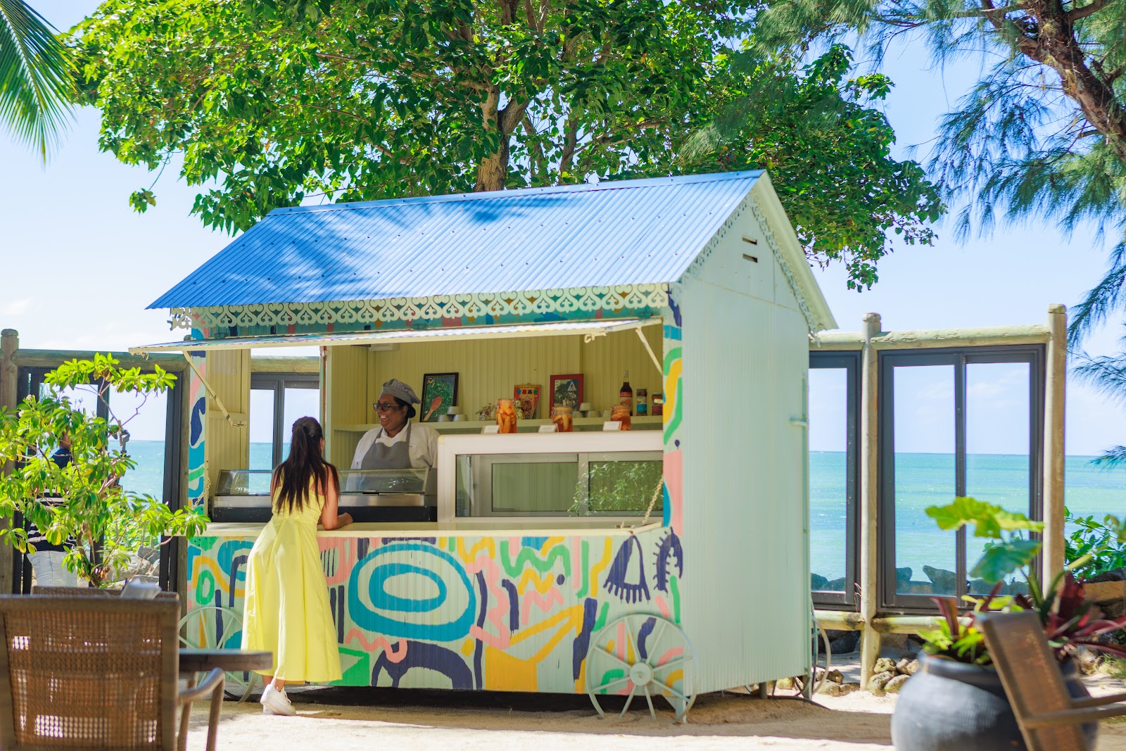 A colorful beachfront food kiosk at Lagoon Attitude Hotel with a server inside and a guest in a yellow dress, turquoise ocean visible beyond.
