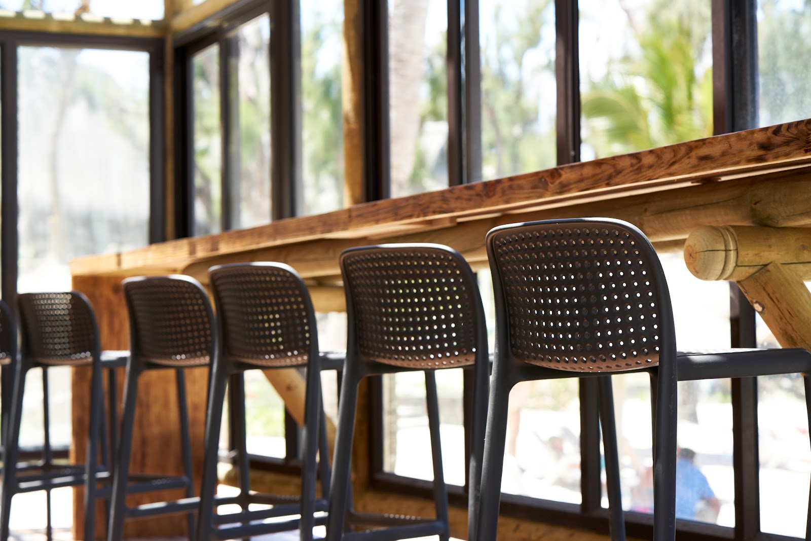 Black perforated bar stools line a wooden counter at Lagoon Attitude Hotel, with large windows and palm trees visible in the background.
