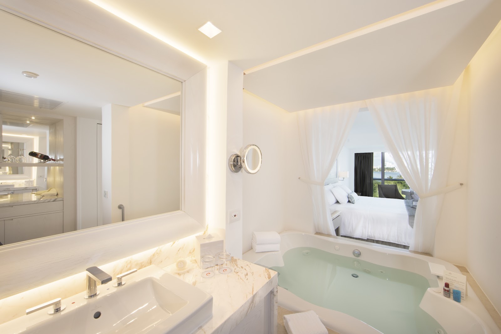 Bright bathroom with soaking tub, illuminated mirror, sink, and view into bedroom with white canopy bed at Le Blanc Spa Resort Cancun.