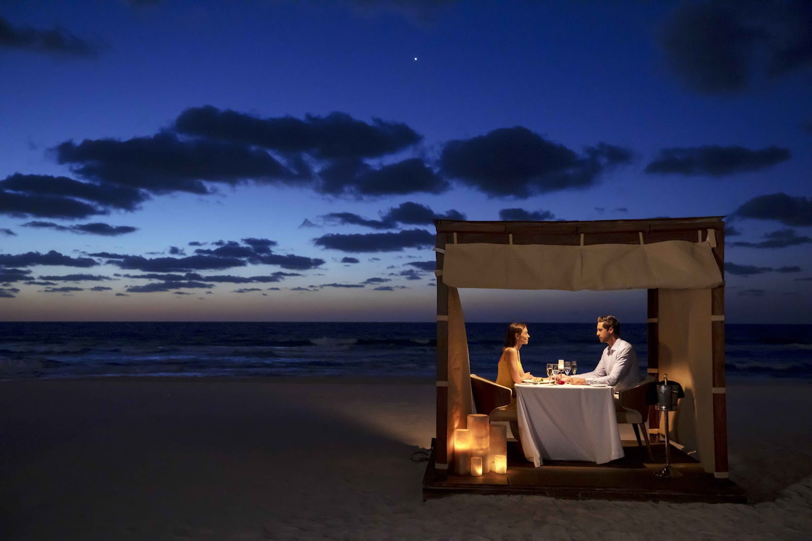 Couple dining at candlelit table beneath wooden cabana on beach at dusk, ocean and twilight sky behind them.