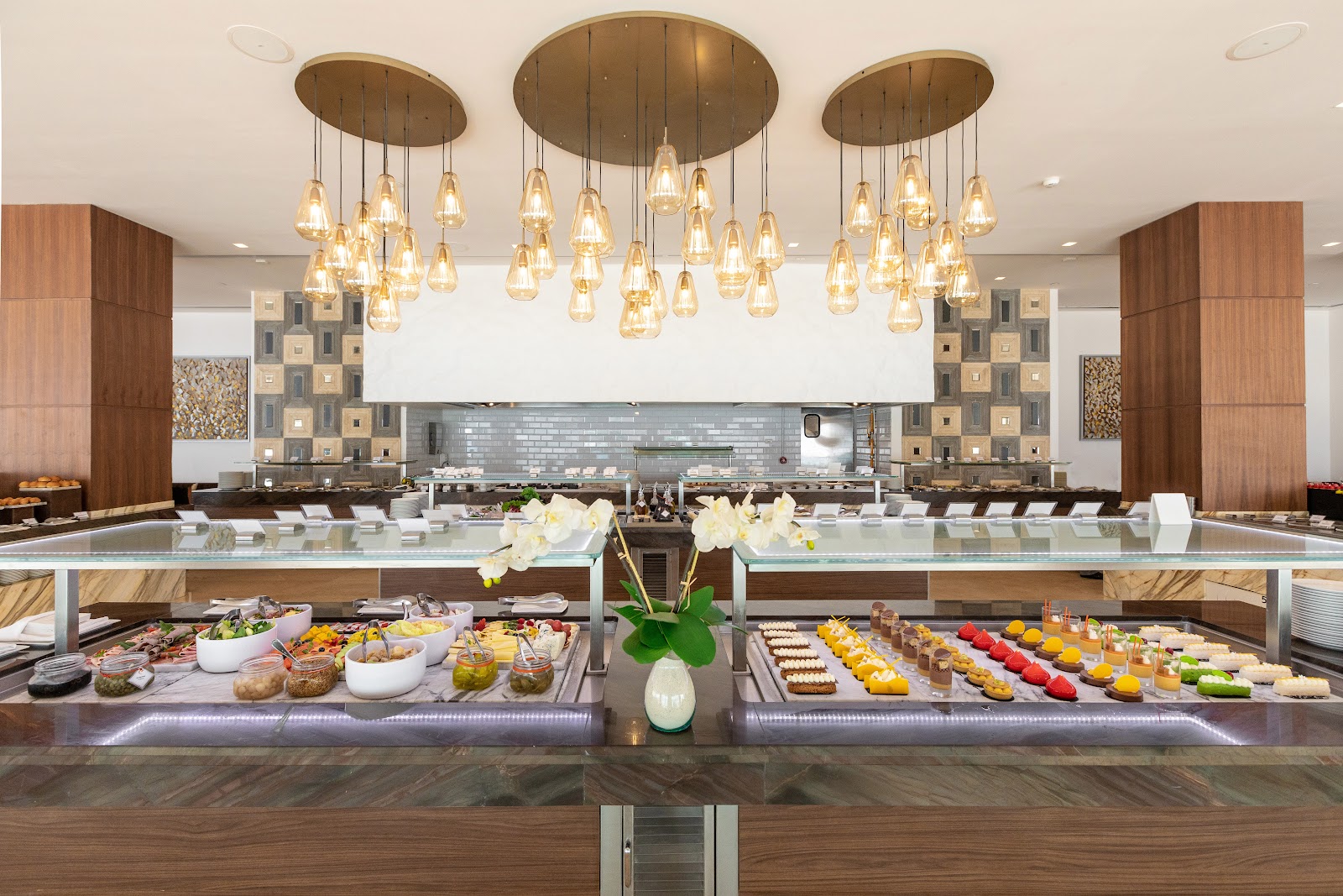 Modern buffet station with glass pendant chandeliers, marble countertops displaying pastries and salads, white orchid centerpiece, and open kitchen at Le Blanc Spa Resort Cancun.