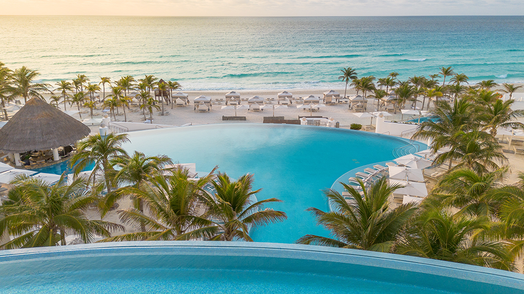 Curved pool in foreground overlooks Le Blanc Spa Resort Cancun's main pool, beach with lounge chairs, palm trees, and turquoise Caribbean Sea.