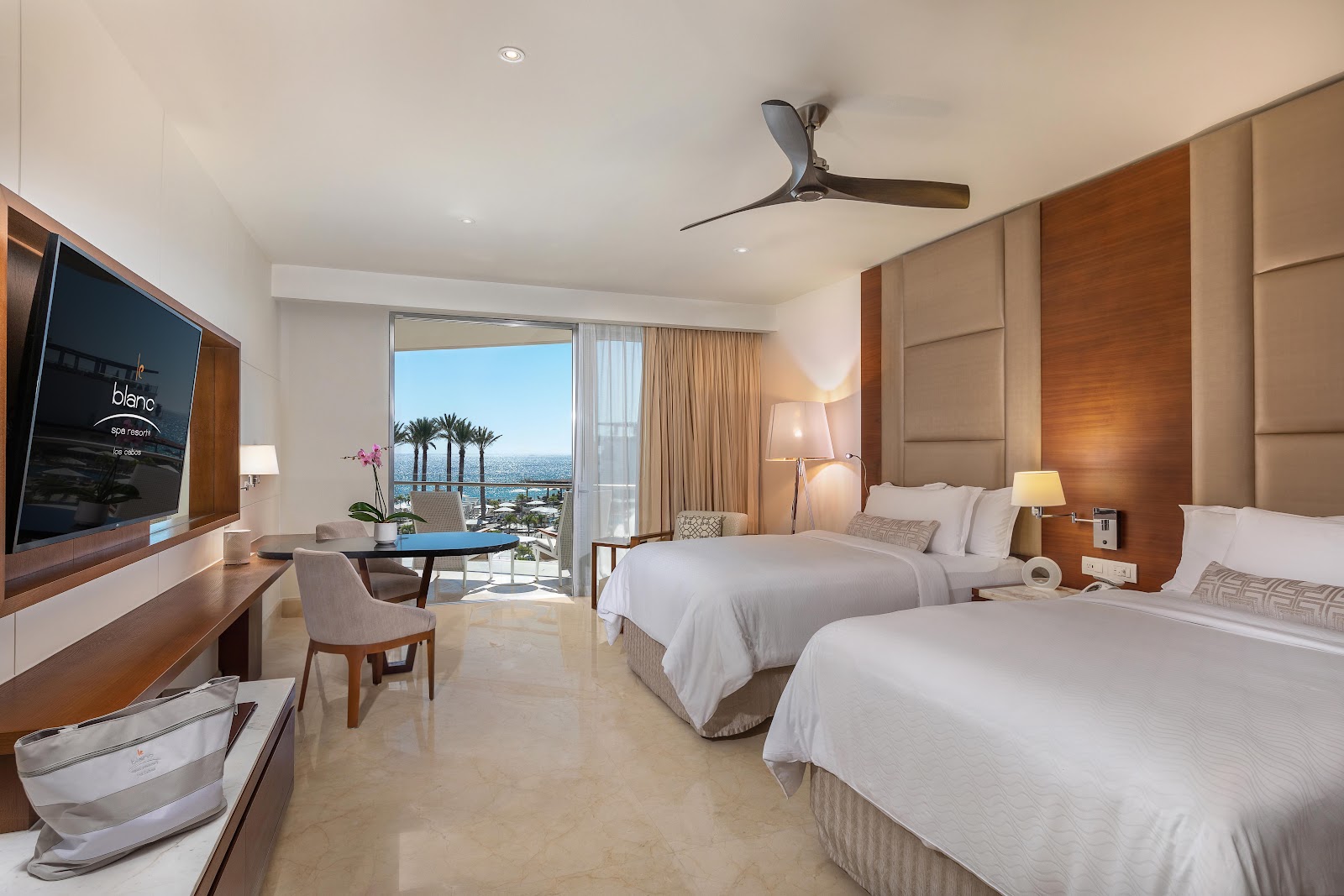 Resort guest room with two queen beds, wood paneling, flat-screen TV, and ocean view from balcony with palm trees visible.