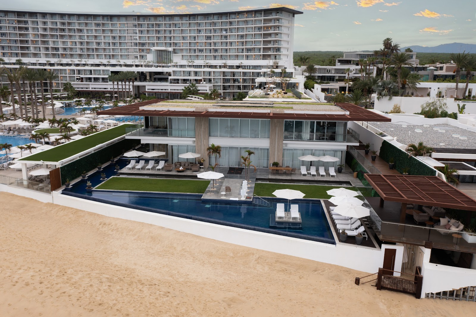 Le Blanc Spa Resort Los Cabos beachfront with modern buildings, infinity pool, lounge areas, and desert mountains in the distance.
