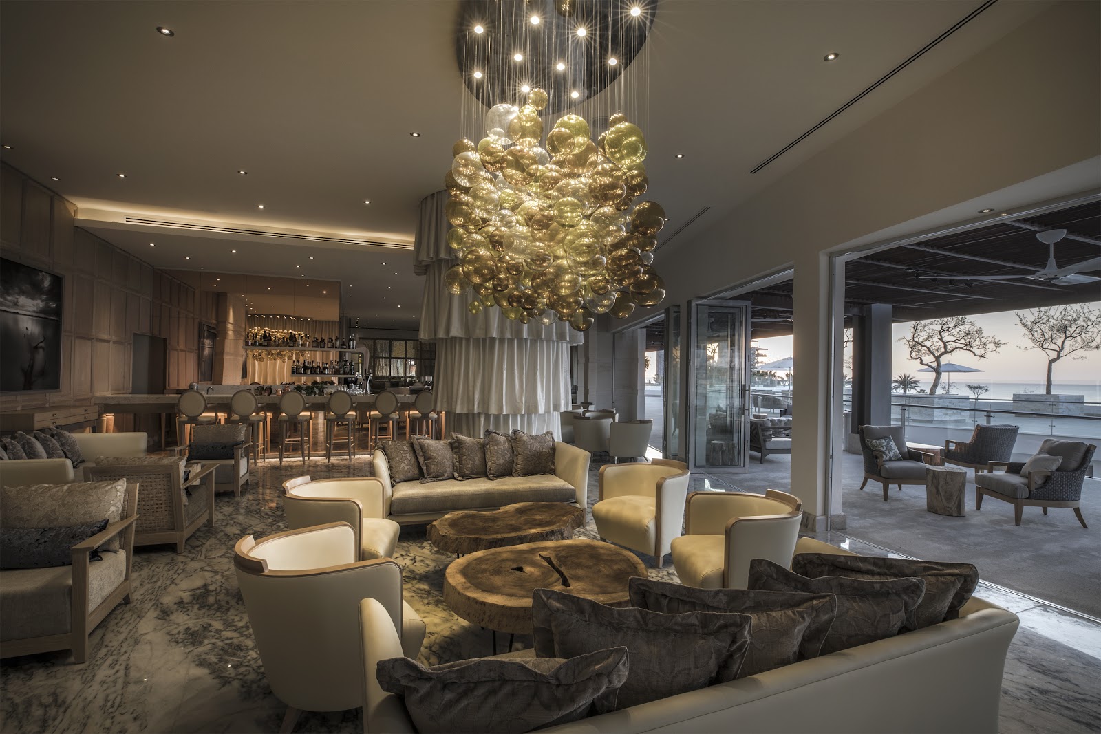 Upscale lobby lounge with cream and charcoal seating, a dangling golden glass chandelier, and ocean views through floor-to-ceiling windows at Le Blanc Spa Resort Los Cabos.