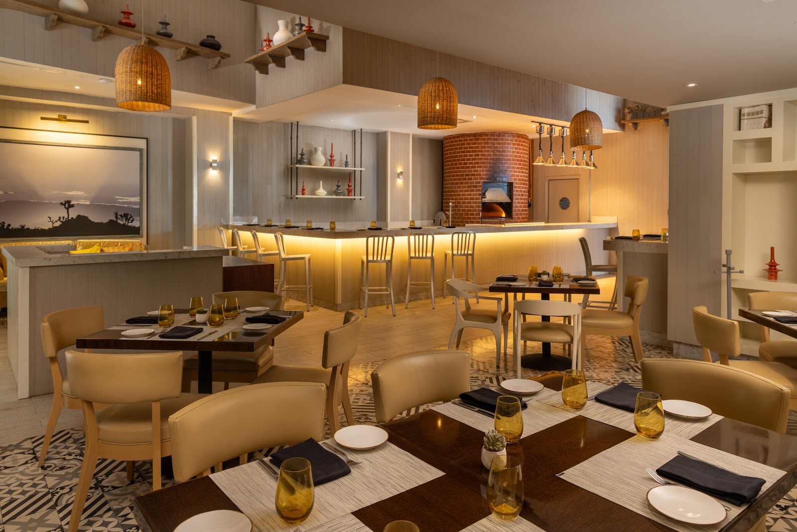Restaurant dining room with cream leather chairs, dark tables set with place settings, a brick pizza oven, and a bar with woven pendant lights at Le Blanc Spa Resort Los Cabos.