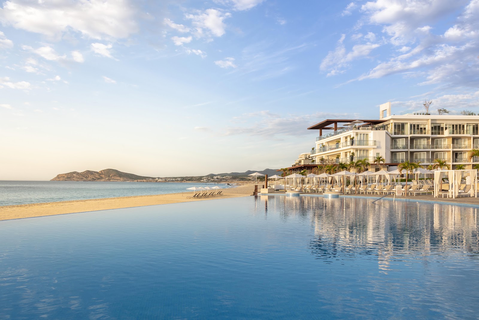 Beachfront resort building with infinity pool overlooking sandy beach and distant mountains under partly cloudy sky.