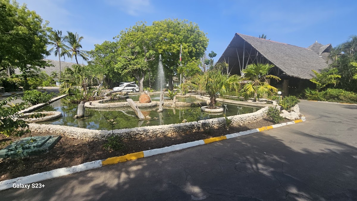 Leopard Beach Resort & Spa entrance drive with a circular pond, fountain, tropical palms, and thatched-roof building under clear sky.