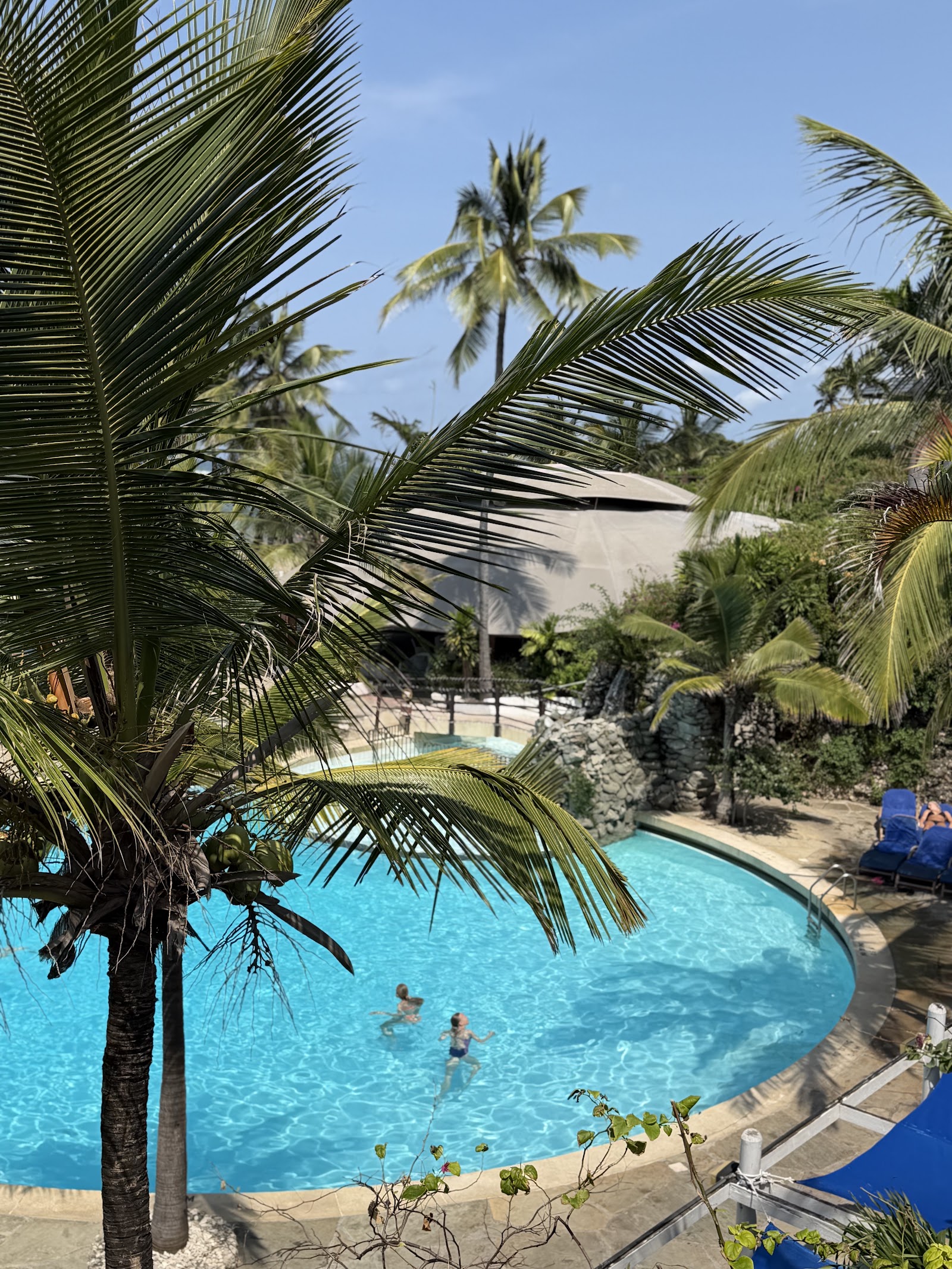 Palm trees frame two blue pools on a sandy beach with the Indian Ocean beyond and a wooden pier extending into turquoise water.