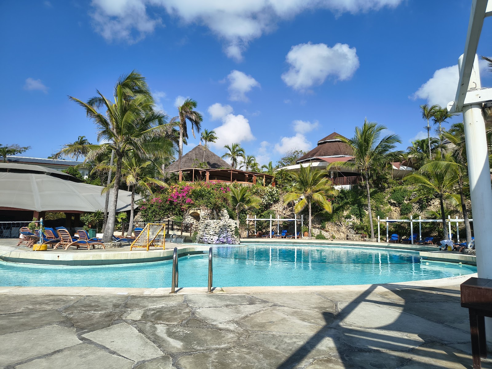 Resort pool with turquoise water surrounded by palm trees, thatched-roof buildings, and lounge chairs under a clear blue sky.