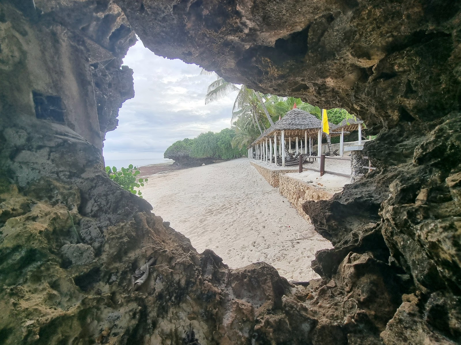 View through a rock cave opening to a sandy beach with thatched-roof bungalows, palm trees, and mangrove forest at Leopard Beach Resort & Spa.