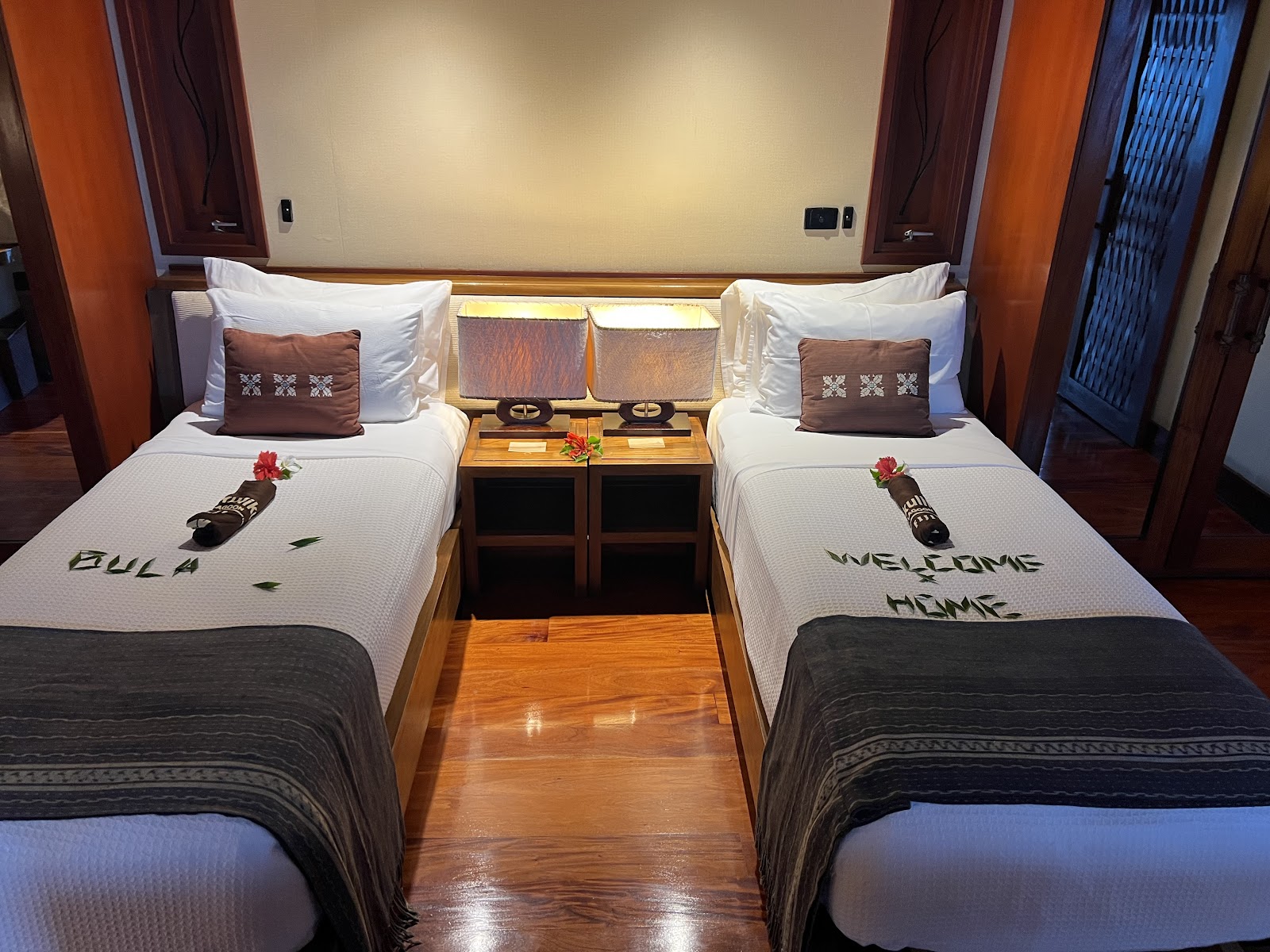 Twin beds with white linens and welcome messages sit in a modern Likuliku Lagoon Resort room with wood floors and warm lighting.