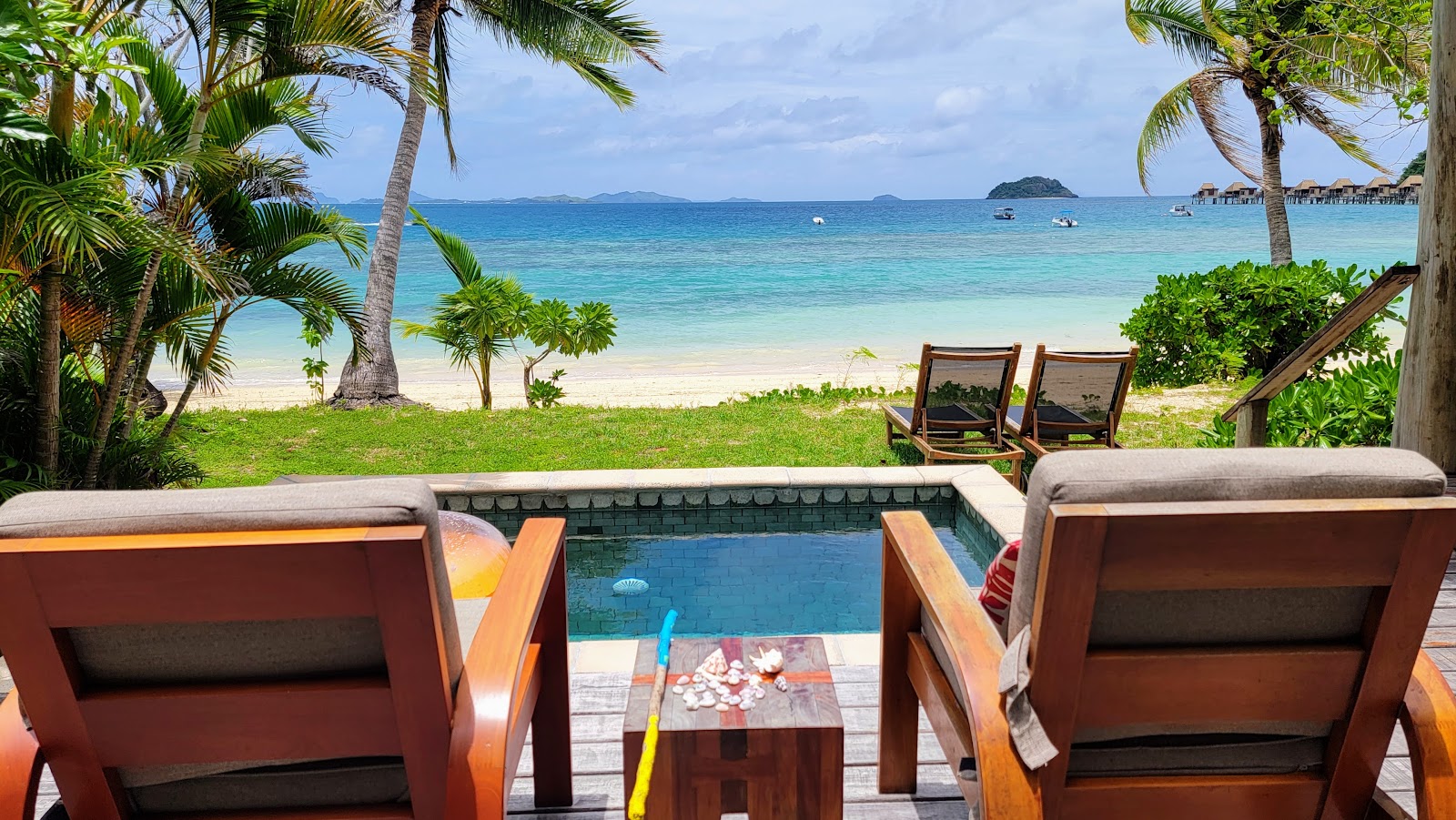 Wooden lounge chairs face a private pool and turquoise lagoon at Likuliku Lagoon Resort, with palm trees and sandy beach beyond.