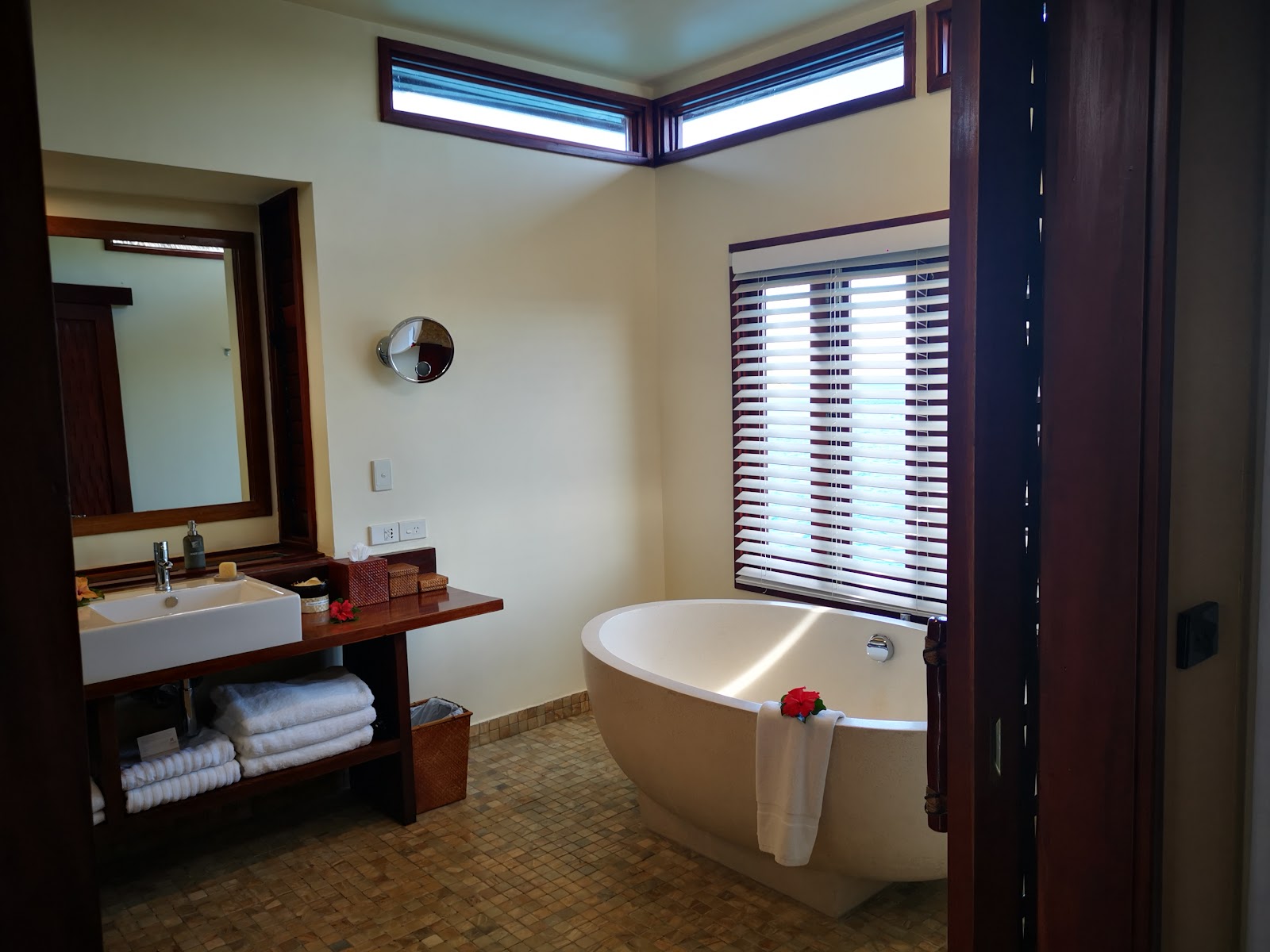 Resort bathroom with sunken soaking tub, wooden vanity with white sink, striped shutters, and folded towels on shelf.