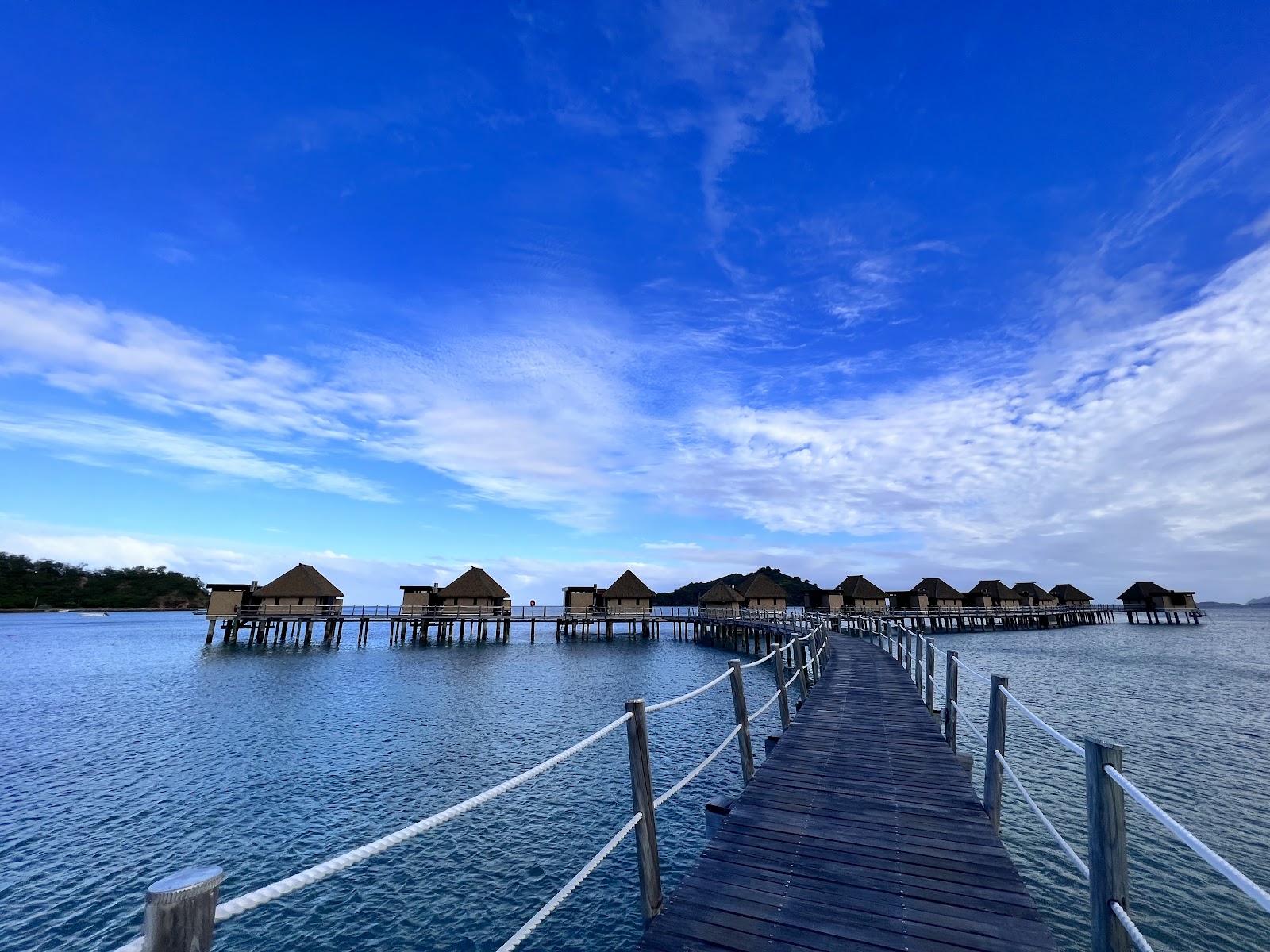 Wooden dock extending into turquoise lagoon toward thatched-roof overwater bungalows at Likuliku Lagoon Resort under clear blue sky.