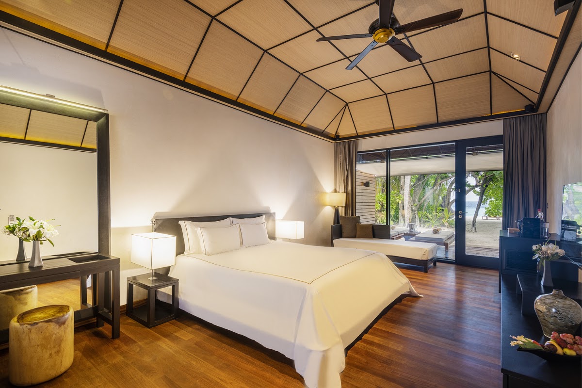 Modern resort bedroom with wooden ceiling, queen bed with cream linens, dark furniture, and glass doors opening to tropical beach views.