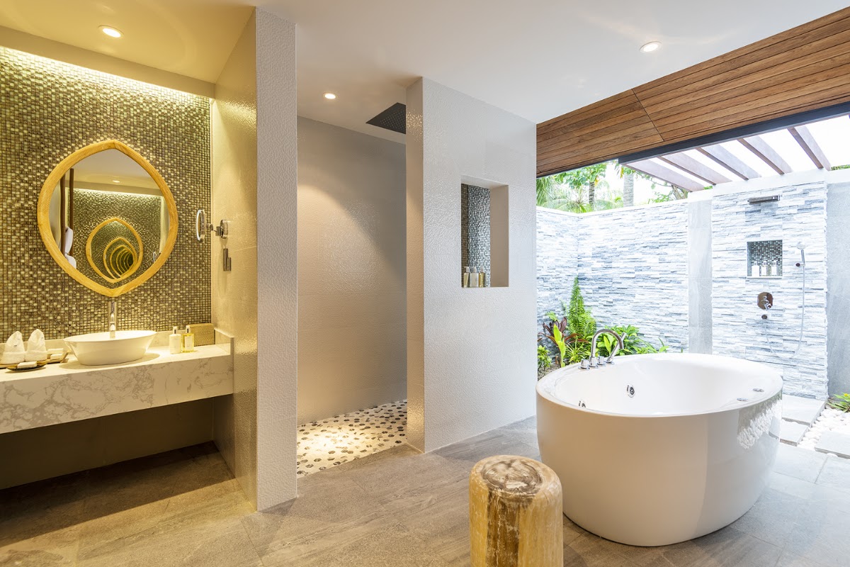 Modern bathroom with round soaking tub, yellow mosaic tile accent wall, circular mirror, and outdoor shower area with stone wall and tropical plants.