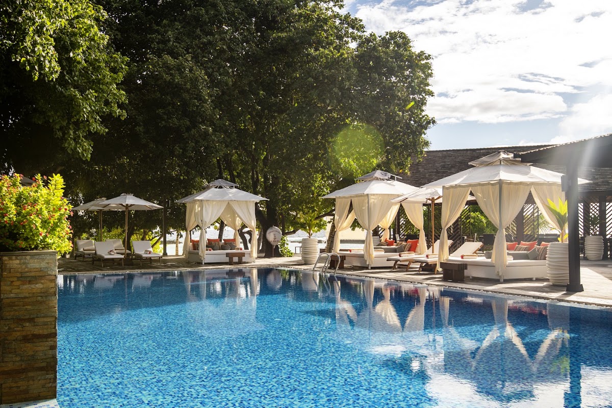 Large blue pool surrounded by cream-colored cabanas and umbrellas with lounge chairs, framed by dense green trees.