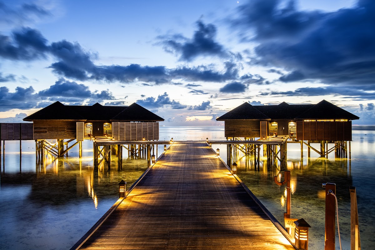Lit wooden boardwalk stretches toward two illuminated overwater bungalows at dusk under a dramatic cloudy sky.