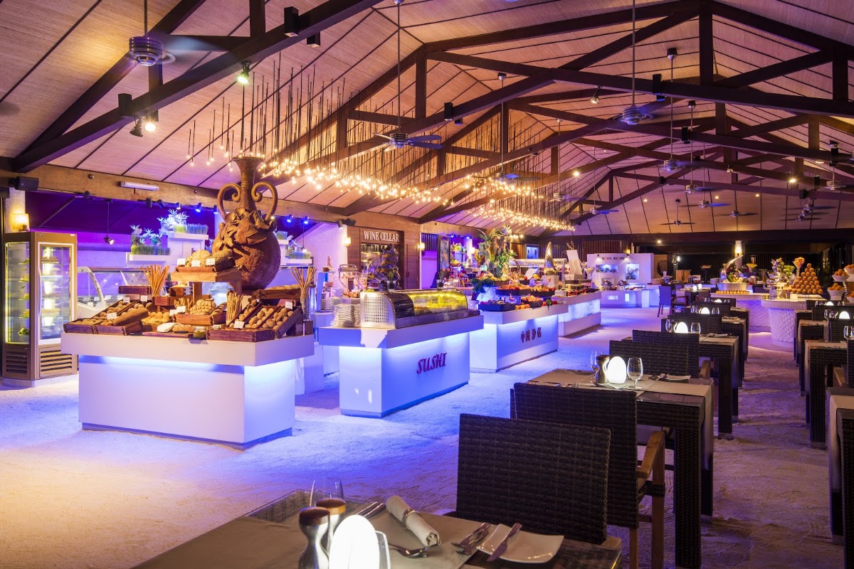 Spacious indoor dining area at Lily Beach Resort with illuminated buffet stations, decorative gold sculptures, string lights, and purple accent lighting throughout the high-ceilinged space.