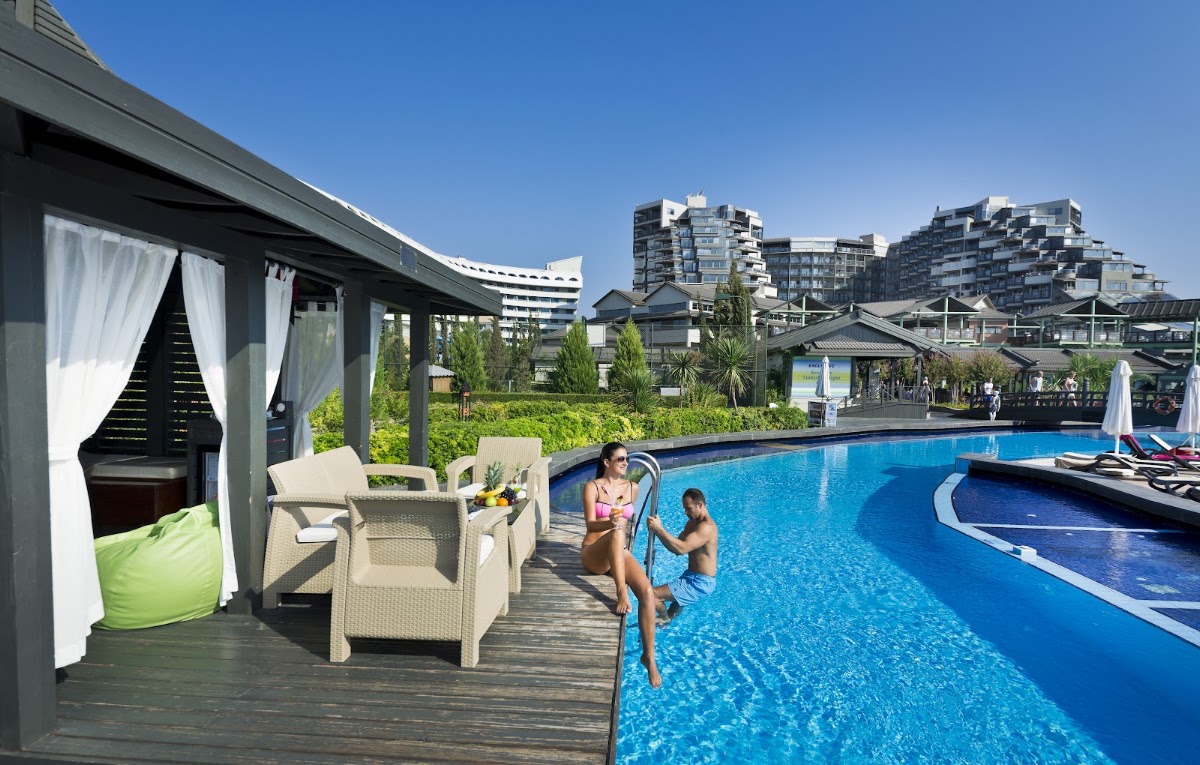 Two guests enter a bright blue pool beside a cabana with white curtains and cream loungers on a wooden deck, with resort buildings in the background.